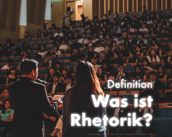 Ein Mann und eine Fau stehen an einem Rednerpult vor einem vollen Hörsaal. Darüber steht: "Was ist Rhetorik? Definition" | © 2022 Claus R. Kullak | Wan San Yip / Unsplash | crk-resrhetorica.de